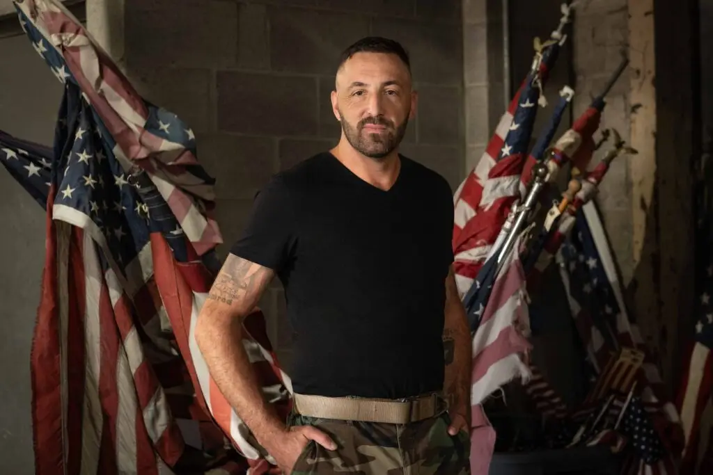 A man in a black t-shirt and camouflage pants stands with his hands on his hips in front of several American flags in a dimly lit, industrial-looking space.