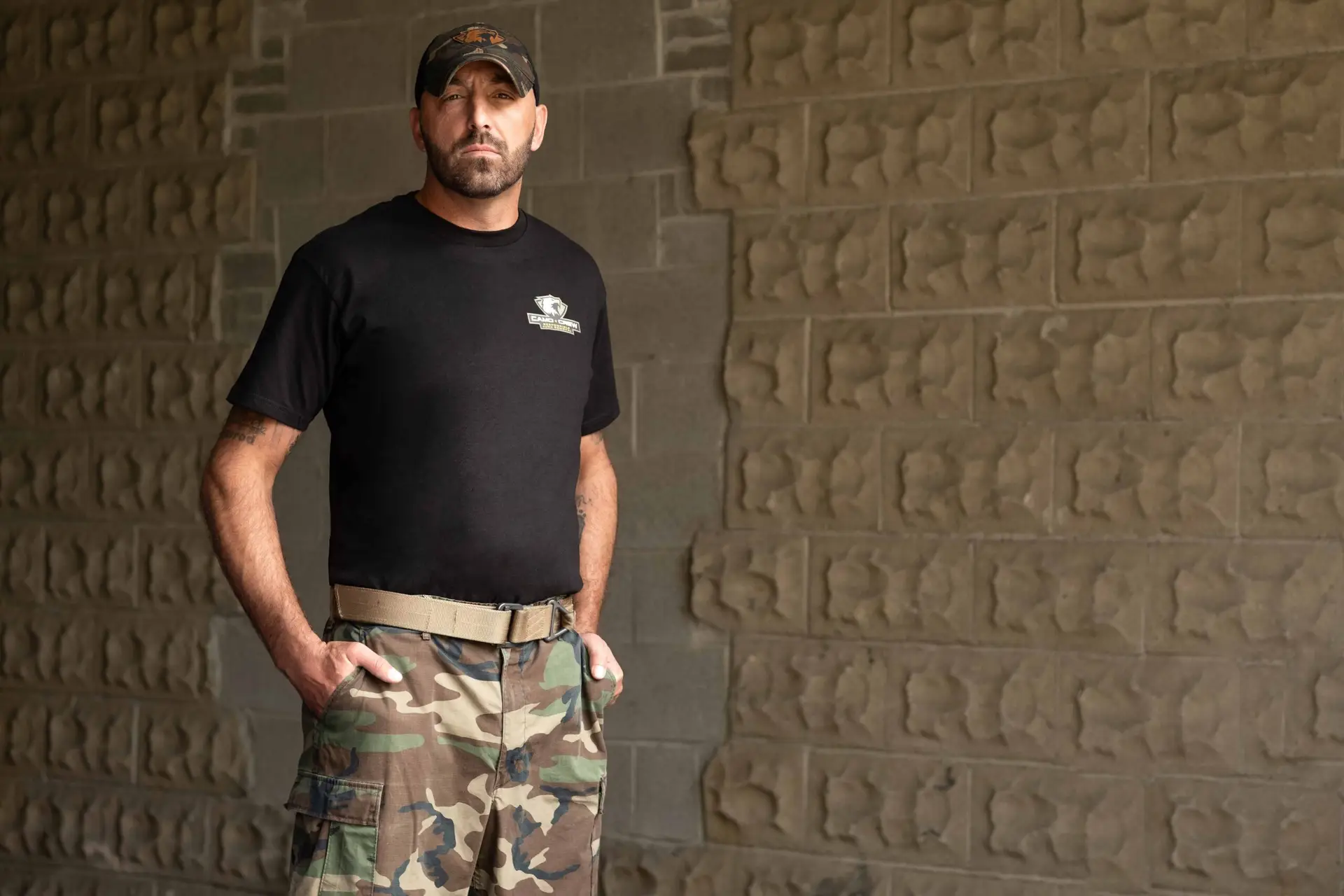 A man wearing a black t-shirt, camouflage pants, and a baseball cap stands with his hands in his pockets against a stone wall.