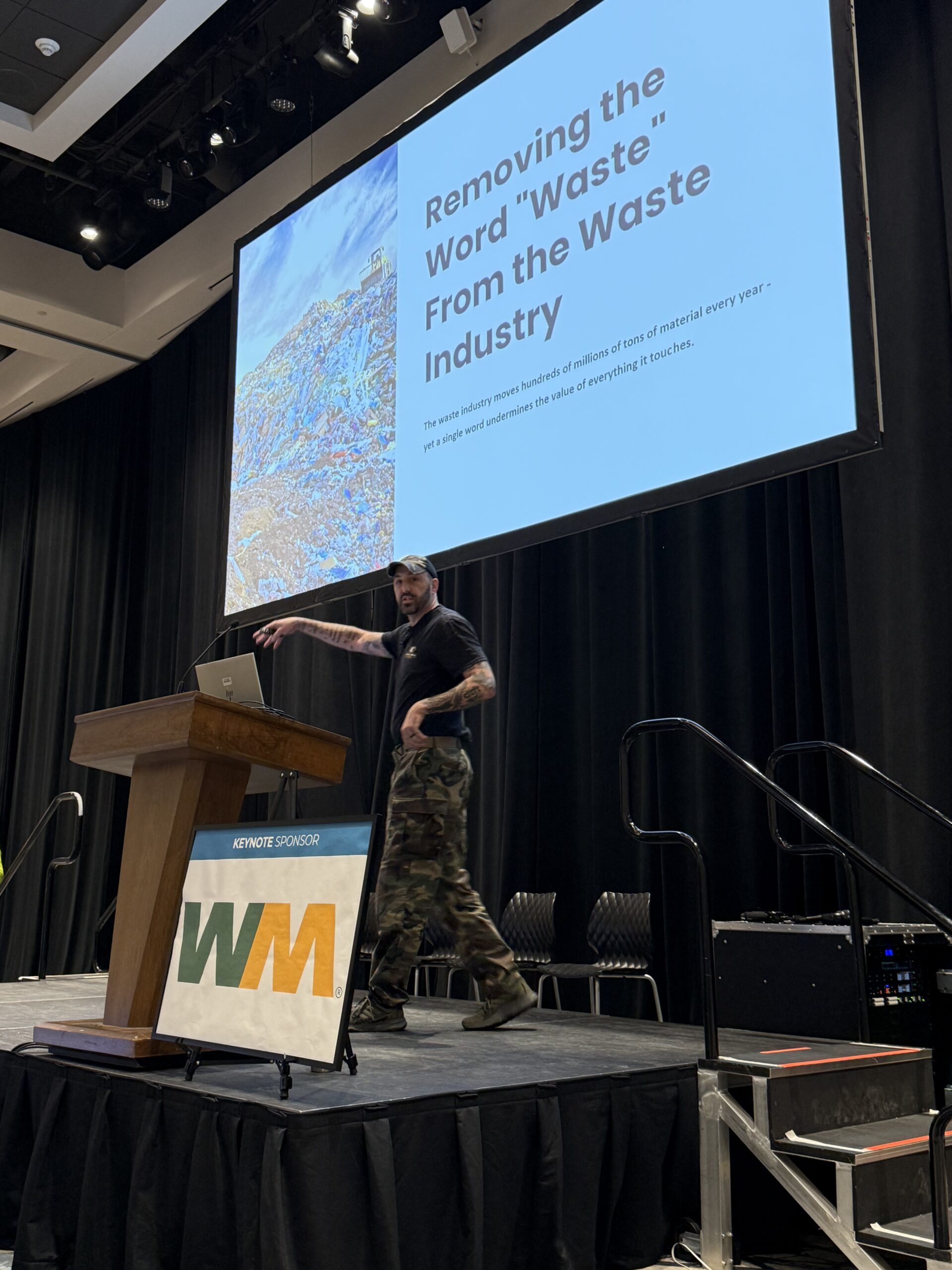 A man in camouflage pants speaks at a podium on stage. Behind him, a large screen reads, Removing the Word Waste From the Waste Industry. A WM sponsor sign is in front of the podium.
