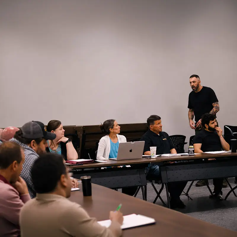 A group of people sit around tables in a meeting room, listening attentively to a man standing and speaking. Laptops, notebooks, and coffee cups are on the tables. The atmosphere appears focused and collaborative.